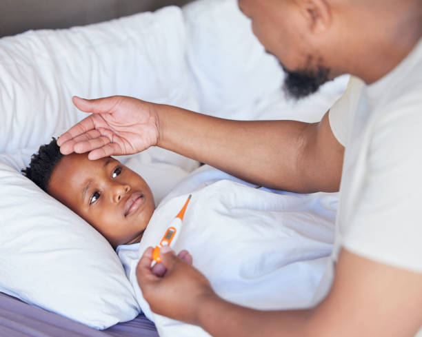 sick little boy in bed while his father uses a thermometer to check his temperature. black single parent feeling son's forehead. african american child feeling ill while his father checks fever - child fever illness thermometer imagens e fotografias de stock