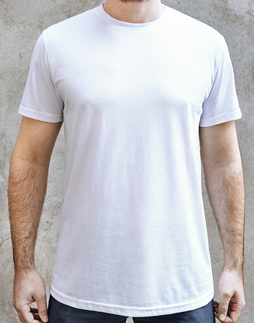 Young man in an empty white casual t-shirt. Front view close up on a background of light gray concrete wall. Design and layout of men's t-shirt for printing.