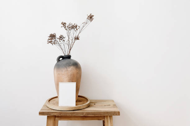 Elegant minimal stationery mockup. Blank greeting card, invitation. Rustic clay vase, pitcher with dry grass, hypericum flowers on old wooden stool. White wall background. Empty copy space. Interior.