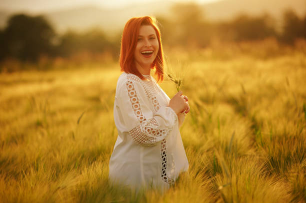 Redhead woman with big smile looking in the camera while holding crop in her hands . She is on a wheat field on a sunny summer day somewhere in Romania