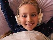 Blonde child girl in headphones learning language listening to music podcast with smartphone online in bed at home.