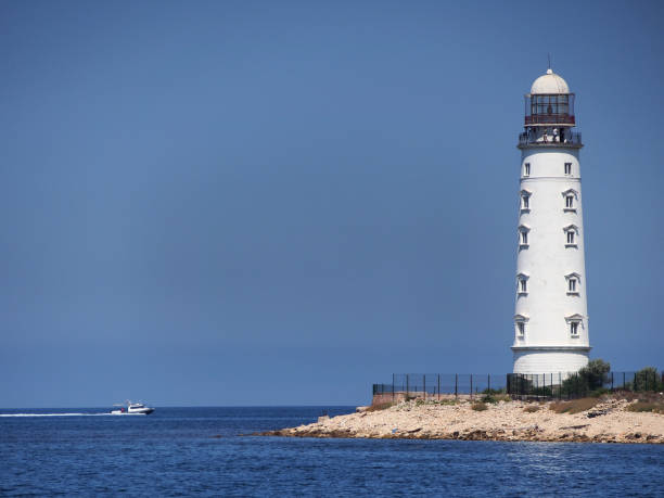 Lighthouse on the shore overgrown with sparse vegetation, against the blue sky.