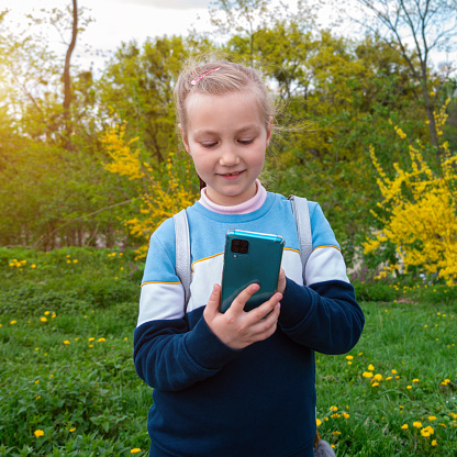 Adorable child girl chatting talking in social media smartphone learning language reading books outdoor. Blogger kid with gadget distance school online podcast lesson cell phone in park yellow flowers