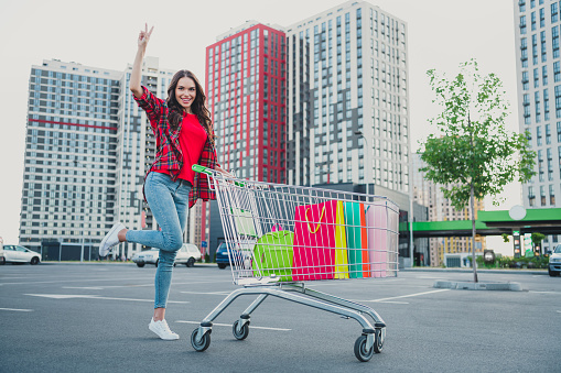 Full length body size view of attractive trendy cheerful funky girl carrying shop cart buying goods showing v-sign on parking outdoors.