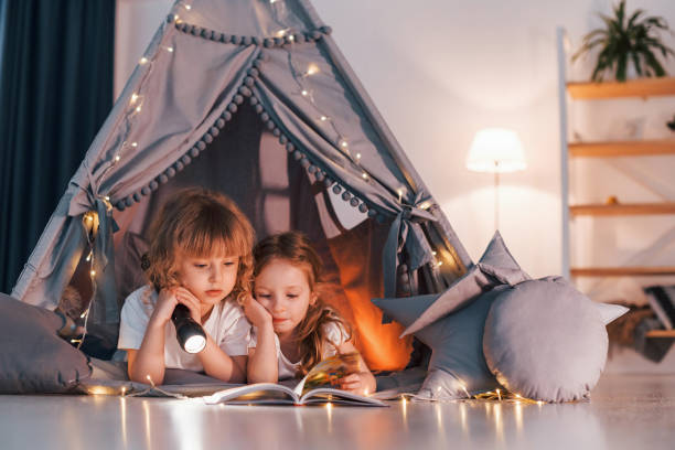 kładzenie się i używanie latarki do czytania książki. dwie małe dziewczynki są razem w namiocie w pokoju domowym - domestic tent zdjęcia i obrazy z banku zdjęć
