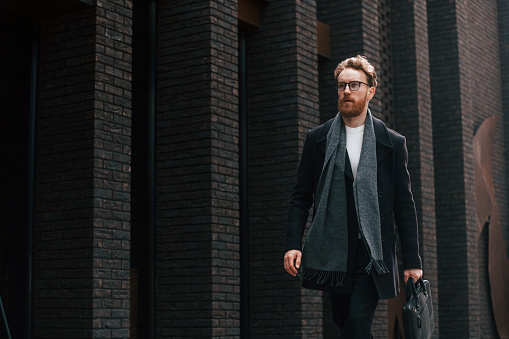 Walking near building. Stylish man with beard and in glasses is outdoors.