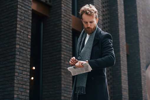 Holding newspaper in the hand. Stylish man with beard is outdoors near building.