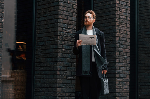 With newspaper in hands. Stylish man with beard and in glasses is outdoors near building.