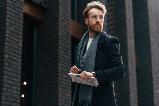 Holding newspaper in the hand. Stylish man with beard is outdoors near building.