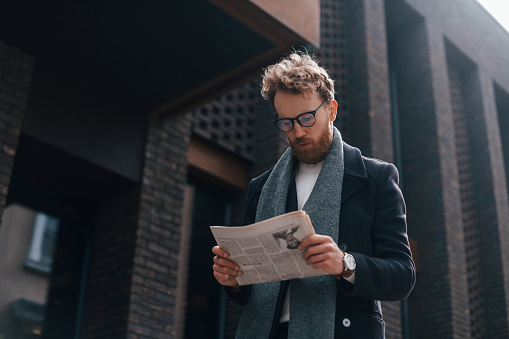 With newspaper in hands. Stylish man with beard and in glasses is outdoors near building.