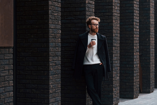 Standing with cup of coffee. Stylish man with beard and in glasses is outdoors near building.