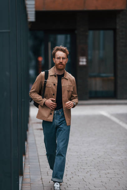 istock Front view. Stylish man with beard in khaki colored jacket and in jeans is outdoors near building 1398103507