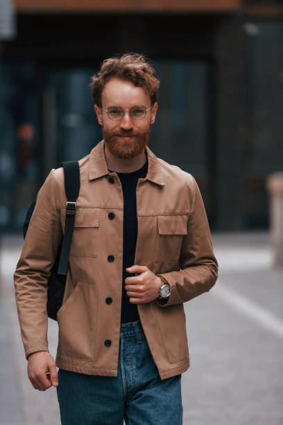 istock Front view. Stylish man with beard in khaki colored jacket and in jeans is outdoors near building 1398103550