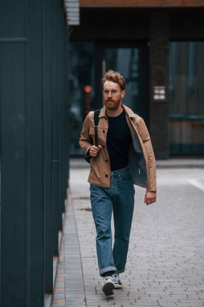 istock Front view. Stylish man with beard in khaki colored jacket and in jeans is outdoors near building 1398103568