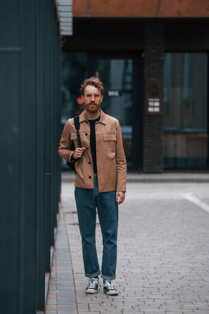 istock Front view. Stylish man with beard in khaki colored jacket and in jeans is outdoors near building 1398103569