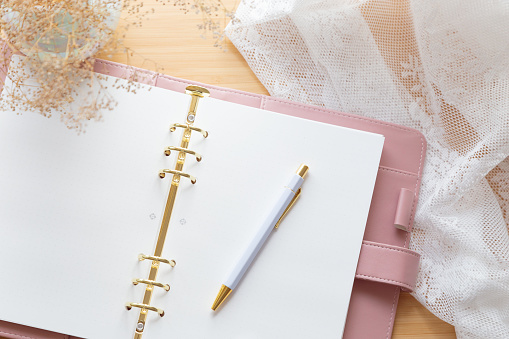 Flat lay, top view of a pastel pink planner, dried flower bouquet and stationery on a wooden desk. Chic workspace, home office, creative writing, wedding planning.