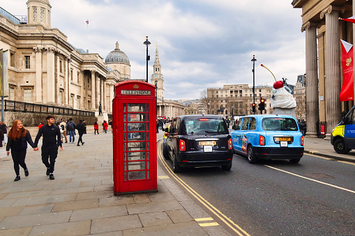 London, United Kingdom - March 6 2022: a back london taxi cab passes a red british phone booth with people passing and the national gallery in the background