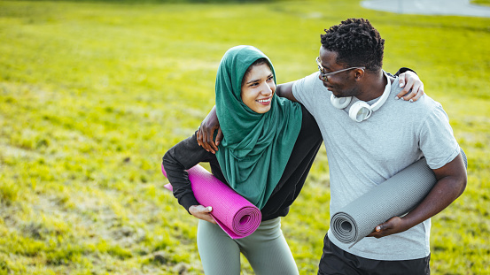 Beautiful positive multicultural couple on sport clothes holding yoga mat in hands while standing outdoors. Young people training regularly on fresh air. Health and body care concept.