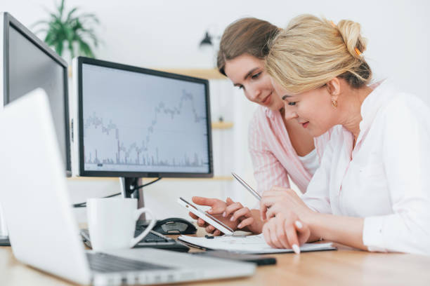 istock Using phone. Two female stock brokers in formal wear is working in the office by pc 1398113437