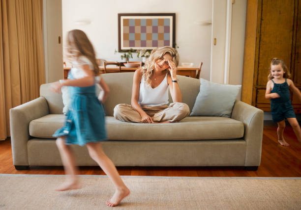 istock Young unhappy caucasian mother suffering from a headache with her daughters running and playing in the lounge at home. Little siblings being playful together while their mom is stressed	sitting on the couch. Woman upset while her children play 1398114641