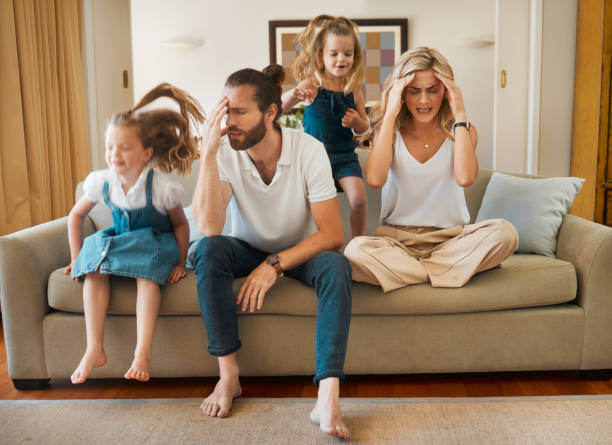 istock Young caucasian mother and father suffering a headache with their daughters playing jumping on the couch in the lounge at home. Little sisters being playful while their parents are stressed. Woman and man tired sitting on the couch 1398114643
