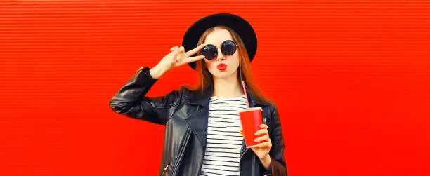 Portrait of stylish young woman model posing with cup of juice blowing her lips wearing black round hat, leather biker rock jacket on red background, blank copy space for advertising text