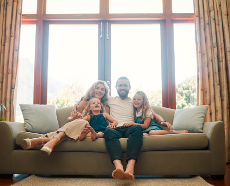 Young happy content caucasian family holding a cardboard as a roof covering them sitting on the floor at home. Cheerful little girls bonding with their mother and father. Loving parents spending time with their cute daughters