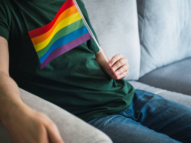 istock Unrecognizable man with copy space and with gay pride day flag sitting on sofa watching tv. Concept person hiding their sexuality 1398120501