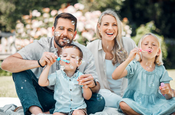 familia caucásica sonriente soplando pompas de jabón por diversión mientras se relajan juntos en el parque o jardín afuera en un día soleado. padres amorosos que se unen con niños juguetones que disfrutan de una infancia feliz y despreocupada - bubble wand bubble child playful fotografías e imágenes de stock