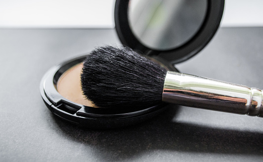 A black makeup brush next to an open box with a mirror and powder puff and pink powder. Black background.