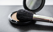 A black makeup brush next to an open box with a mirror and powder puff and pink powder. Black background