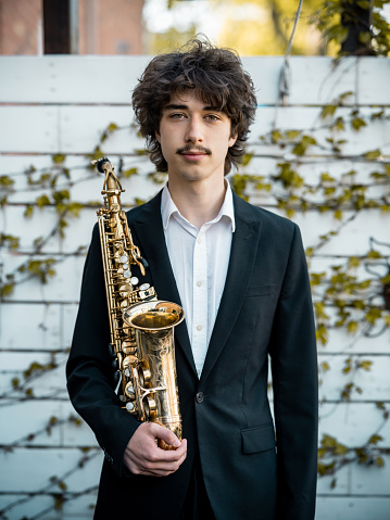 Outdoor Portrait of young male saxophone player. Young Caucasian man with messy hair and moustache, wearing white dress shirt and black coat. Outdoors during the day.