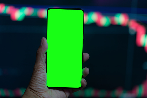 Financial Analyst Using Smartphone with Green Screen Chroma Key Mock Up Template and Working on Multi-Monitor Workstation with Stocks Charts. Businessman Works in Investment Bank