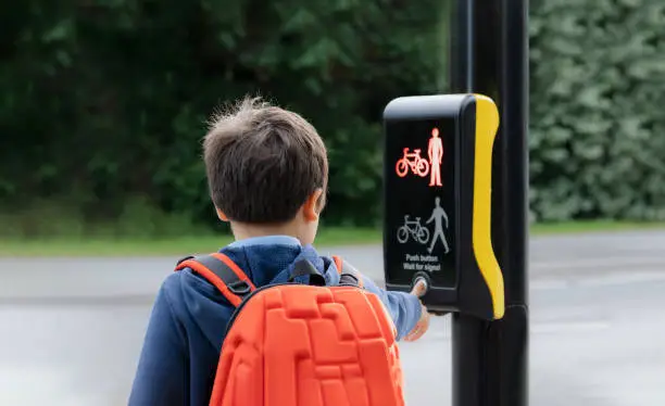 Photo of Rear view portrait School kid pressing a button at traffic lights on pedestrian crossing on way to school. Child boy with backpack using traffic signal controlled pedestrian facilities for crossing road.