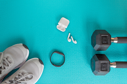image from above of  two 8lb weights, wireless earbuds, headphones, earphones, running shoes and a fitness tracker watch on a turquoise blue yoga mat background
