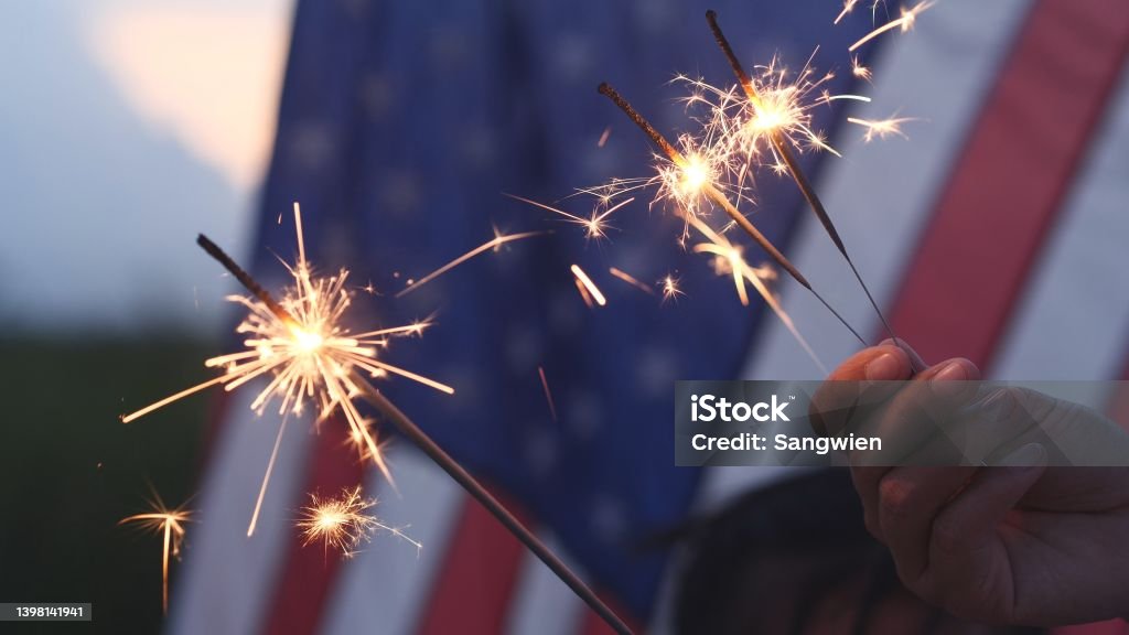 Happy 4th of July Independence Day, Hand holding Sparkler fireworks USA celebration with American flag background. Concept of Fourth of July, Independence Day, Fireworks, Sparkler, Memorial, Veterans Fourth of July Stock Photo