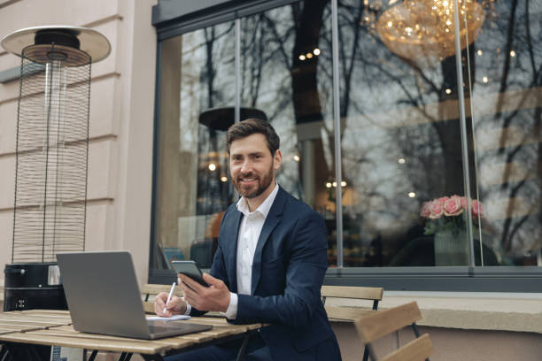 istock Happy businessman with laptop, phone and notebook in cafe, smiling. Taking notes and planning. 1398144593