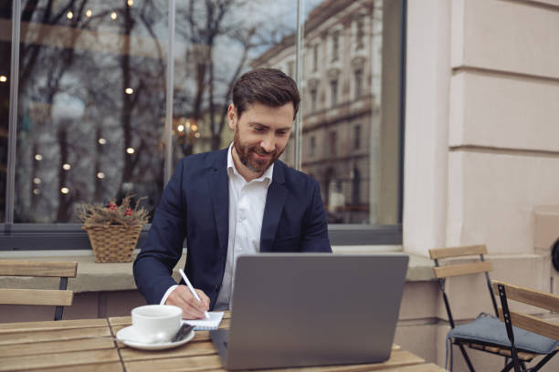 istock Cheerful businessman working at laptop in cafeteria and taking notes in notebook. Coffee break. 1398144594