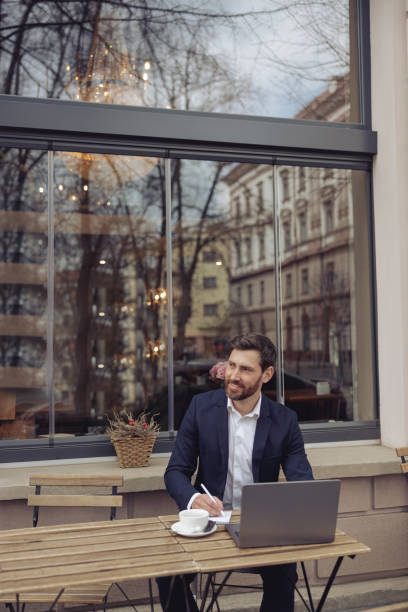 Attractive elegant man taking notes while working at laptop in cafe outside. Coffee break.