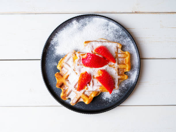 istock Freshly baked Viennese waffles sprinkled with powdered sugar with strawberries lying on a black ceramic plate on a wooden background. Top view 1398148236
