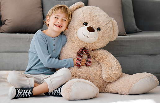 Portrait of one happy little caucasian boy smiling while hugging a big and cosy fluffy teddy bear on the floor in the lounge at home. Adorable kid relaxing and playing with soft stuffed toy alone