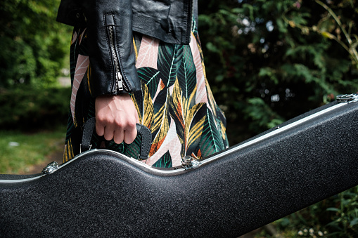 Close up of woman arm holding a guitar case in a park. She is wearing a black leather jacket and a spring dress. Horizontal.
