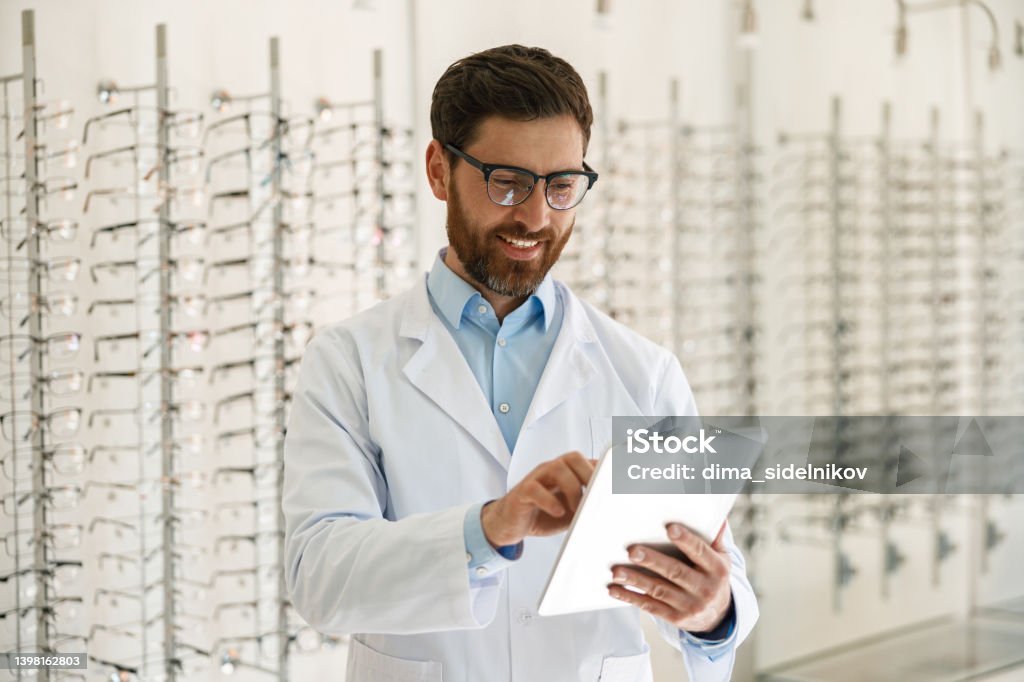 Smiling ophthalmologist working on digital tablet in front of the showcase with eyeglasses Ophthalmologist working on digital tablet in front of the showcase with eyeglasses in optical store Optometrist Stock Photo