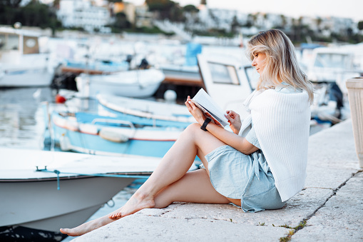 Portrait of young woman walking along embankment with boats and expensive yachts in resort city, sitting on edge of pier and reading book side view. Sea voyage, boat mooring, relaxing leisure.
