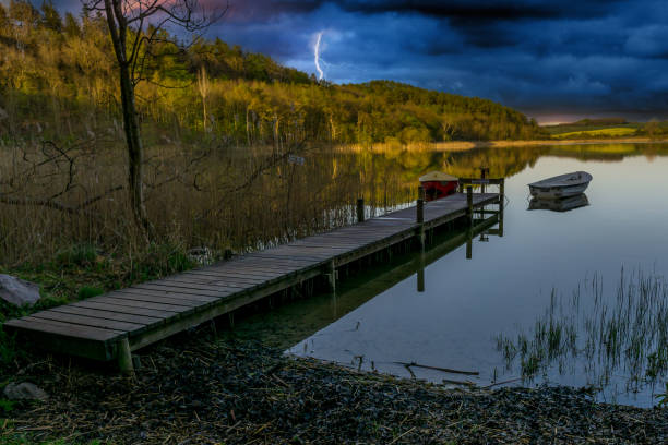 This image captures a serene scene at a calm lake in Bestensee, Dahme-Spreewald, Brandenburg, Deutschland. A wooden dock extends into the lake, providing a perfect viewpoint to appreciate the surrounding forest and the cloudy sky reflected on the water’s surface.
