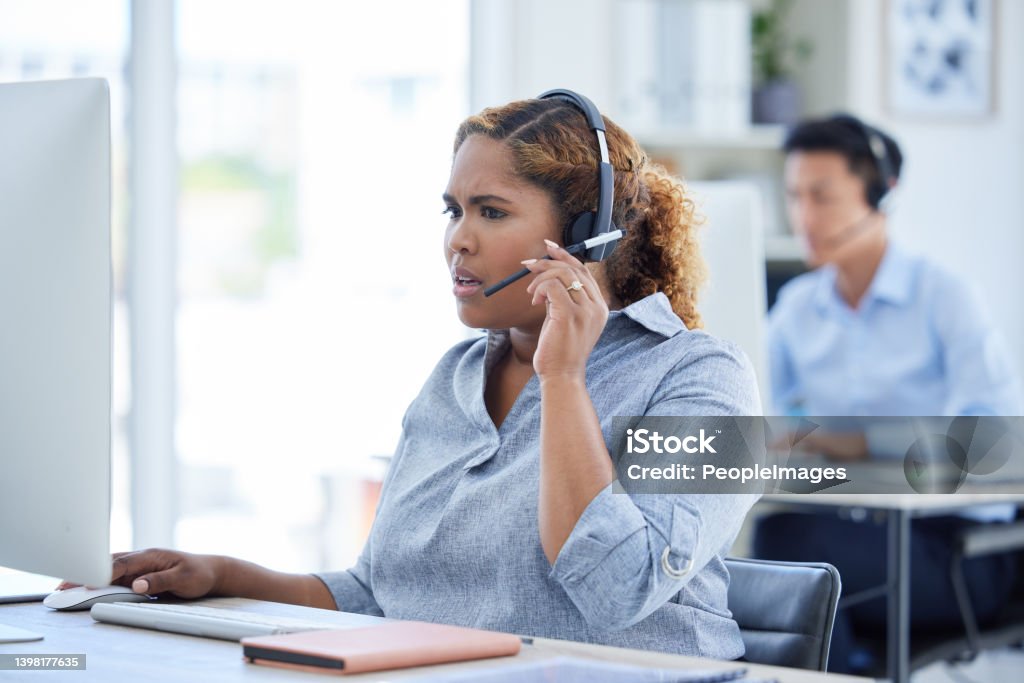 One stressed and confused mixed race call centre telemarketing agent talking on a headset while working on a computer in an office. Young shocked african american female consultant dealing with panic crisis and difficult caller for customer support Call Center Stock Photo