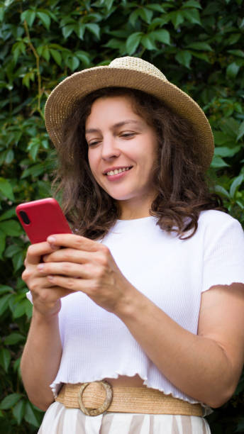 istock Young smiling brunette woman holding mobile phone,using app on smartphone,texting message,browsing standing in green park.Female blogger in summer hat sharing data on social media,scrolling.Vertical 1398178971