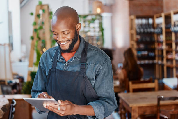 istock Young african american businessman working in a restaurant and  using a digital tablet device. Smiling black man using the the internet to check social media and buy stock. Black entrepreneur at work 1398179492