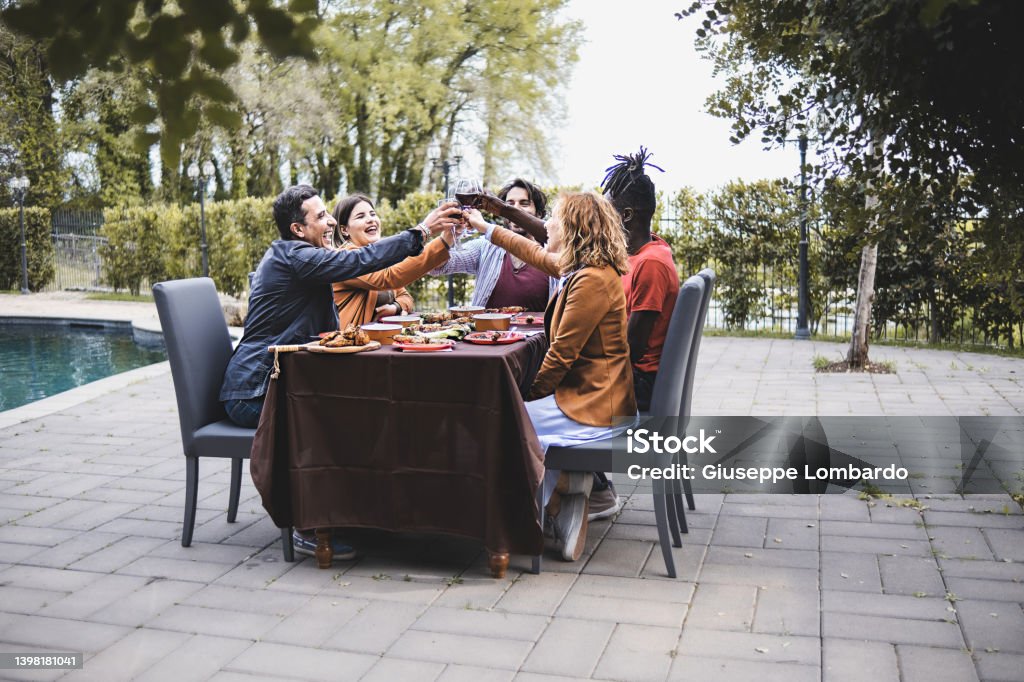 Multiethnic group of friends sitting on a table by the pool celebrating a friendship toasting together - men and women best friends toasting arms up rising wine glasses outdoors Back Yard Stock Photo