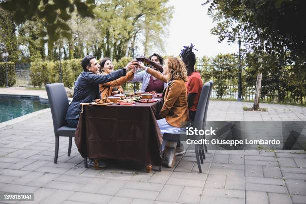 Multiethnic Group Of Friends Sitting On A Table By The Pool Celebrating A Friendship Toasting Together Men And Women Best Friends Toasting Arms Up Rising Wine Glasses Outdoors Stock Photo - Download Image Now